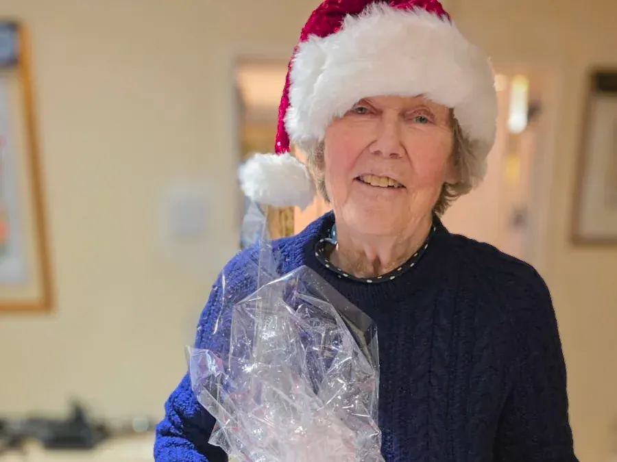 woman wearing a Santa hat and navy jumper holding a cellophane-wrapped Christmas gift indoors.