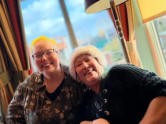 Two women in Christmas hats and jumpers smiling together near a window decorated for the festive season.