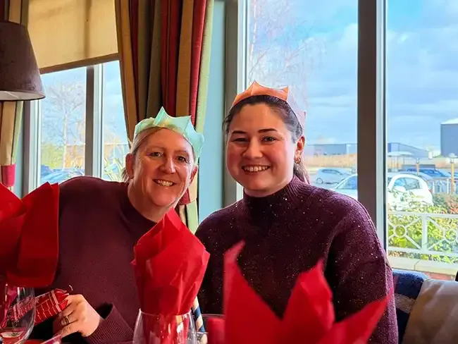 Two women wearing coloured paper crowns smiling at the camera while seated at a restaurant Christmas meal.