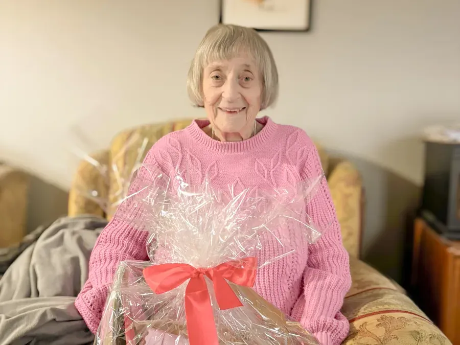woman in a pink jumper sitting on a patterned sofa with a large cellophane-wrapped gift basket tied with a red bow.
