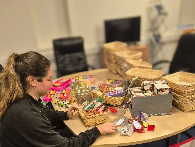 Staff member wrapping a Christmas gift basket in cellophane at a table covered with hampers, cards, and treats.