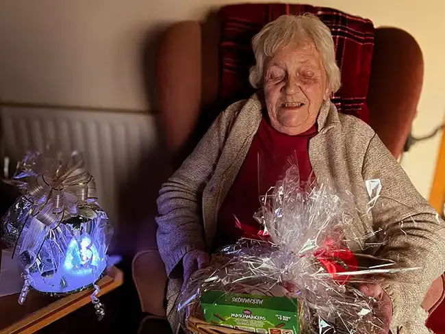 Client sitting in an armchair holding a wrapped Christmas gift basket with a red bow, next to a glowing festive ornament.