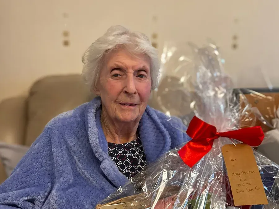 woman in a blue robe sitting on a sofa beside a cellophane-wrapped gift basket with a red bow and handwritten tag.