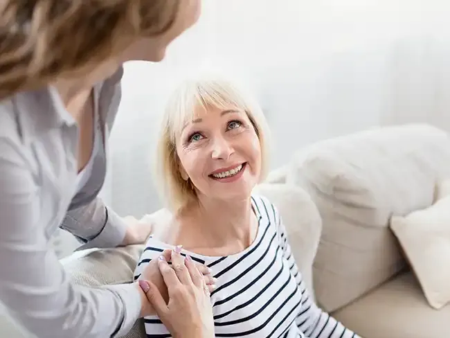 A smiling older woman sitting on a sofa is comforted by a family caregiver holding her shoulder, symbolising how preventative care provides reassurance and support for both the person receiving care and their loved ones.
