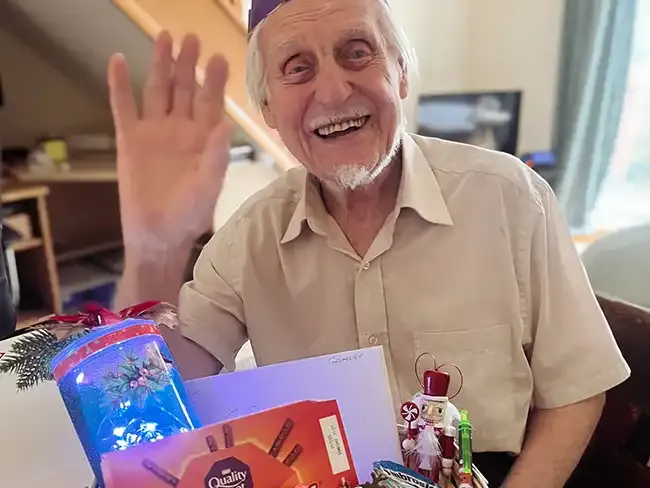 Client waving and smiling with Christmas gifts on the table, including chocolates, a festive light, and a card.