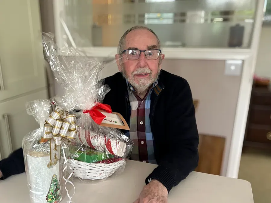 Older man wearing glasses seated at a table with a Christmas gift basket wrapped in cellophane and ribbon.