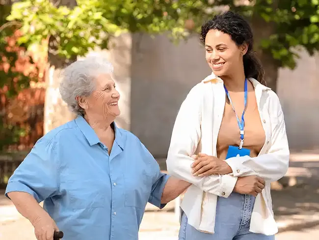 Smiling elderly woman walking outdoors with the support of a young female caregiver, representing independence through preventative home care.