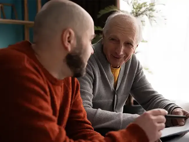 An older man talking with a younger man in a warm indoor setting, discussing winter preparations featured in the Winter Home Safety Checklist.