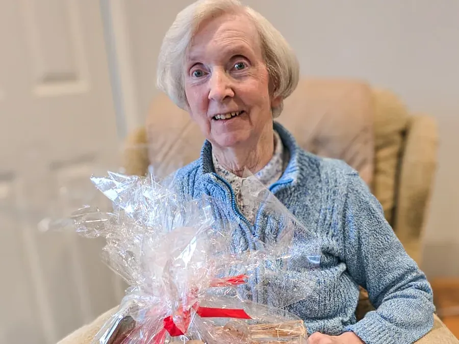 Smiling woman in a light blue cardigan sitting in an armchair with a wrapped Christmas hamper on her lap.