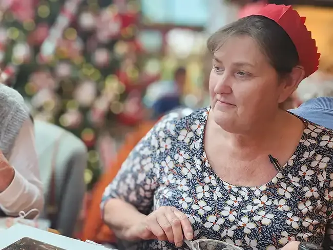 A woman wearing a red paper crown seated at a Christmas lunch table with a decorated tree in the background.