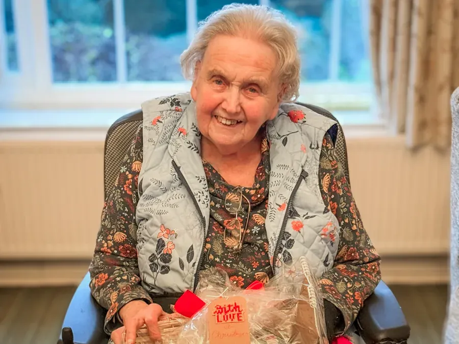 Smiling woman in a floral top and quilted vest seated by a window with a Christmas hamper in front of her.
