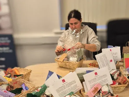 Staff member assembling Christmas hampers at a table with baskets, chocolates, and festive items, with stacks of empty baskets in the background.