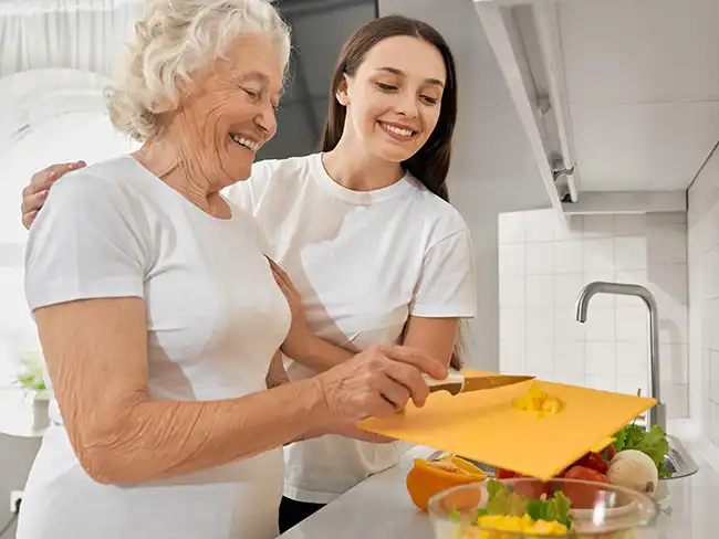 Smiling elderly woman preparing food with the support of a young female carer in a bright kitchen, showing healthy living and preventative home care.