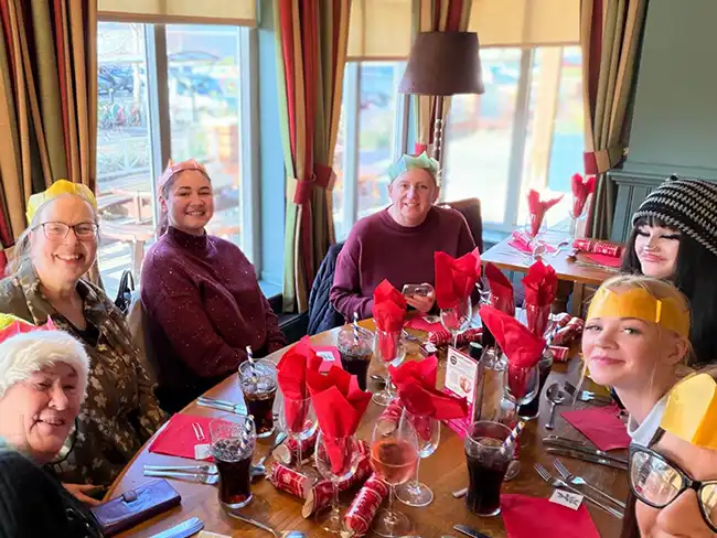 A group of six people sitting together at a Christmas meal table with crackers, red napkins, and drinks, enjoying a festive lunch.