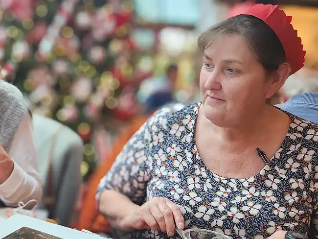 A woman wearing a red paper crown seated at a Christmas lunch table with a decorated tree in the background.