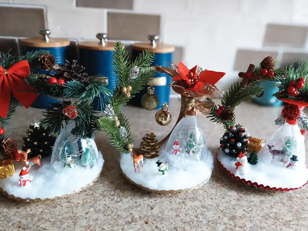 Handmade acrylic Christmas dome displays with snowy bases, mini trees and figures, topped with festive greenery, berries, pinecones and hanging baubles.