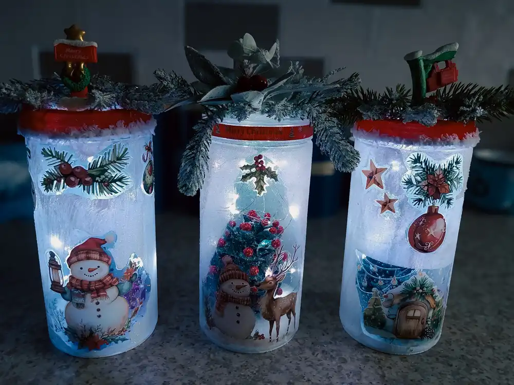 Three LED Christmas jars glowing with snowman, reindeer and festive tree designs, finished with frosted greenery and seasonal toppers.