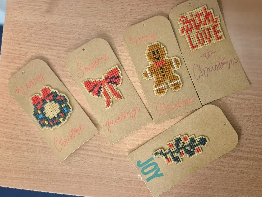 Handmade gift tags with cross-stitched Christmas designs (wreath, bow, gingerbread figure, festive lettering) laid out on a wooden table.
