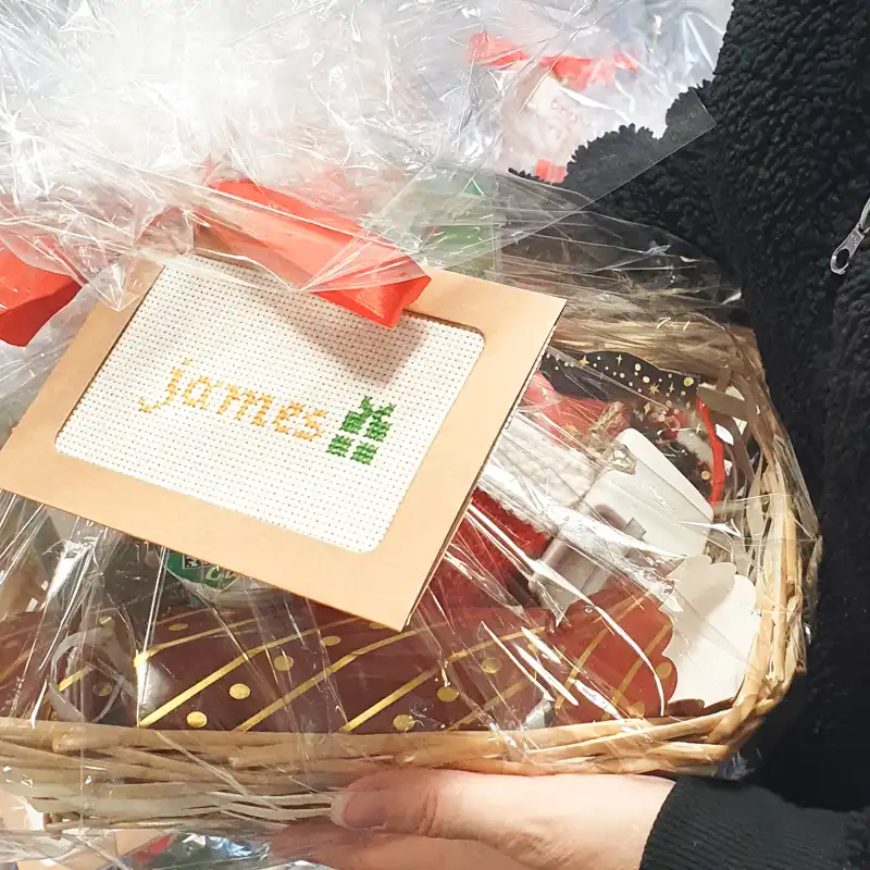 Close-up of a festive client gift basket wrapped in cellophane with a red bow and a personalised ‘Jane’ gift tag, filled with Christmas treats and a winter kit.