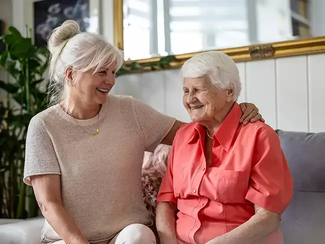 Smiling elderly woman sitting on a sofa with her adult daughter, sharing a warm moment at home &mdash; representing compassionate preventative care and family support.