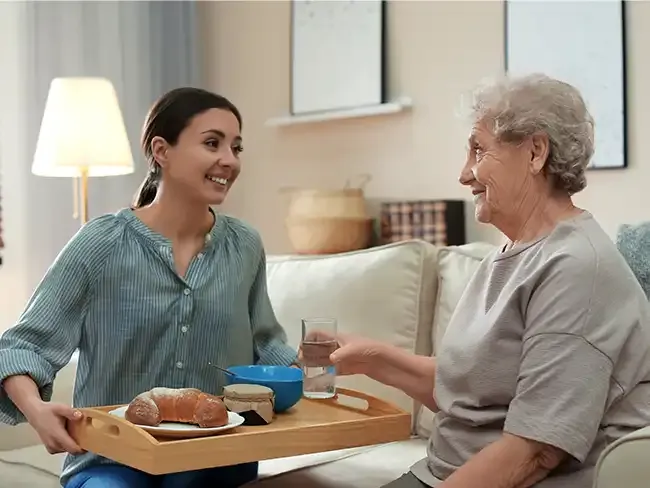 Young caregiver serving breakfast on a tray to an elderly woman sitting on a sofa, both smiling warmly at each other.