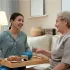 Young caregiver serving breakfast on a tray to an elderly woman sitting on a sofa, both smiling warmly at each other.