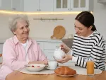 Smiling elderly woman sitting at a kitchen table while a young respite caregiver pours tea into her cup.