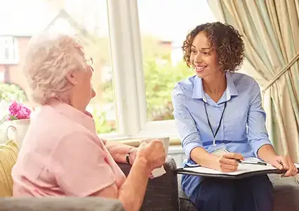 Smiling carer providing elderly care at home, talking with an older woman and offering support and guidance.