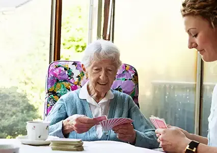 Elderly woman playing cards with a carer at home, enjoying companionship and support through elderly care services.
